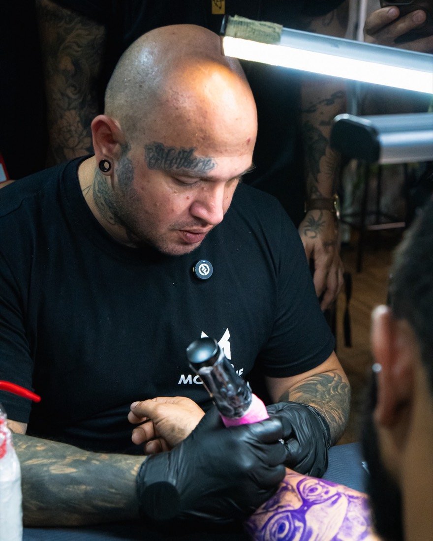 Chico Morbene tatuando o antebraço de um cliente, de camiseta do Grupo Morbene e máquina na mão.